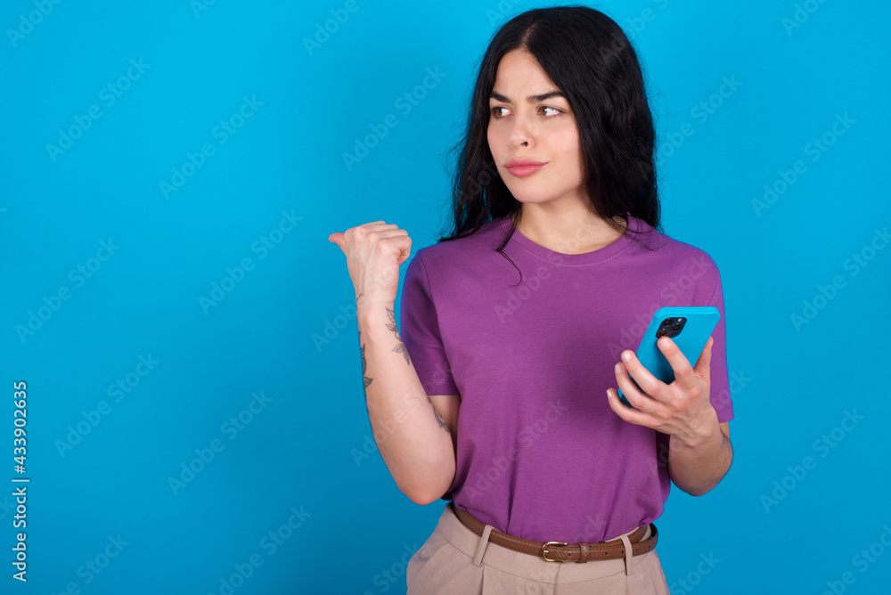 young beautiful tattooed girl wearing blue t-shirt standing against blue background points thumb away and shows blank space aside, holds mobile phone for sending text messages. young beautiful tattooed girl wearing blue t-shirt standing against blue background points thumb away and shows blank space aside, holds mobile phone for sending text messages.