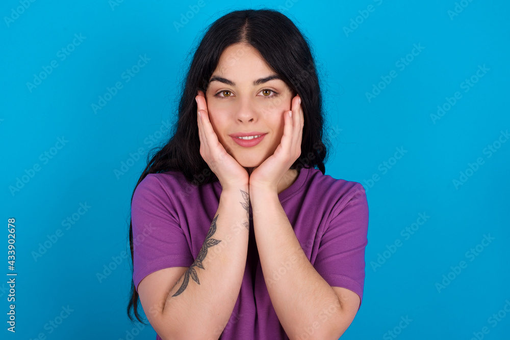 Happy young beautiful tattooed girl wearing blue t-shirt standing against blue background touches both cheeks gently, has tender smile, shows white teeth, gazes positively straightly at camera, Happy young beautiful tattooed girl wearing blue t-shirt standing against blue background touches both cheeks gently, has tender smile, shows white teeth, gazes positively straightly at camera,