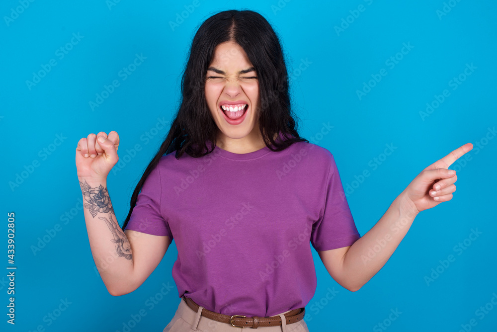 Cheerful young beautiful tattooed girl wearing blue t-shirt standing against blue background showing copy space ad celebrating luck Cheerful young beautiful tattooed girl wearing blue t-shirt standing against blue background showing copy space ad celebrating luck