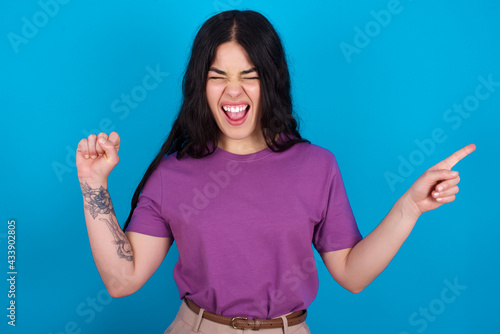 Cheerful young beautiful tattooed girl wearing blue t-shirt standing against blue background showing copy space ad celebrating luck