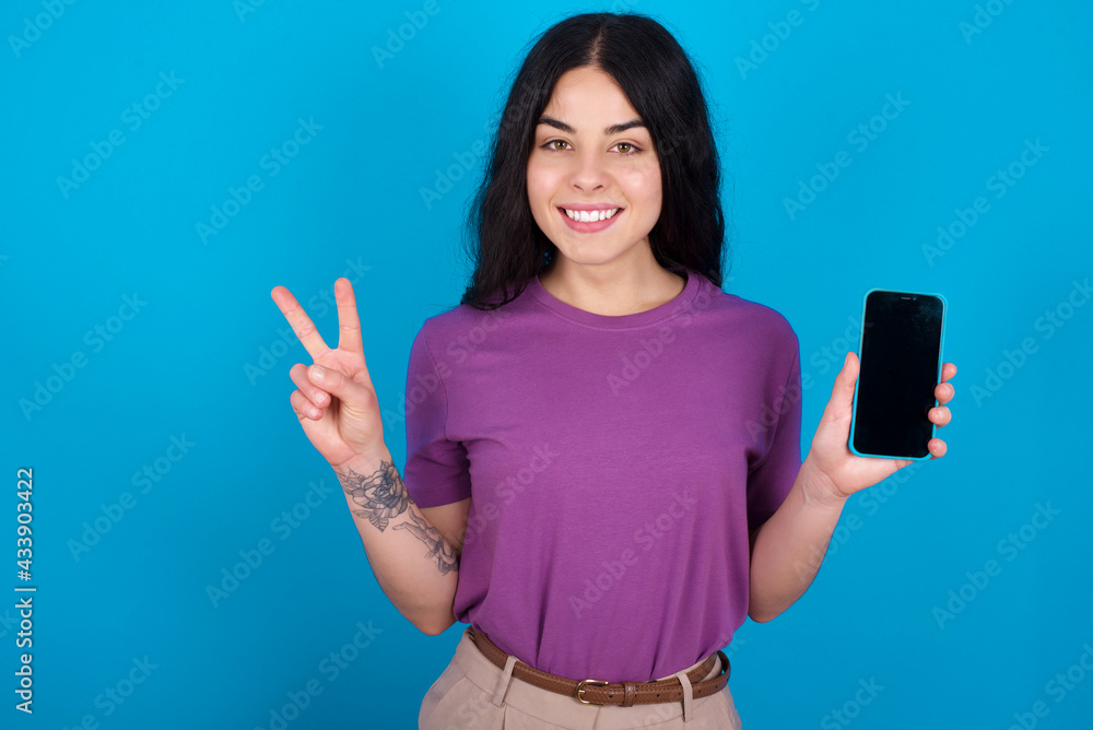 young beautiful tattooed girl wearing purple t-shirt standing against blue background holding modern device showing v-sign young beautiful tattooed girl wearing purple t-shirt standing against blue background holding modern device showing v-sign