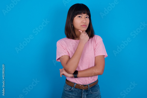 Thoughtful smiling young beautiful asian woman wearing pink t-shirt against blue wall keeps hand under chin, looks directly at camera, listens something with interest. Youth concept.