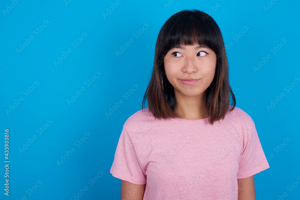 Amazed puzzled young beautiful asian woman wearing pink t-shirt against blue wall, curves lips and has worried look, sees something awful in front. Amazed puzzled young beautiful asian woman wearing pink t-shirt against blue wall, curves lips and has worried look, sees something awful in front.