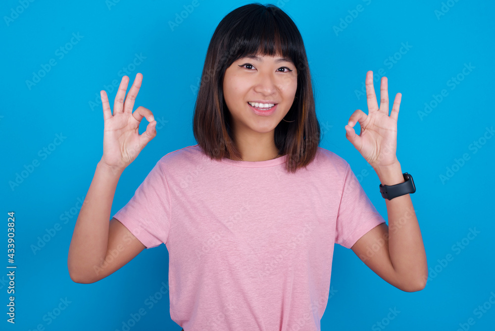 Glad young beautiful asian woman wearing pink t-shirt against blue wall shows ok sign with both hands as expresses approval, has cheerful expression, being optimistic. Glad young beautiful asian woman wearing pink t-shirt against blue wall shows ok sign with both hands as expresses approval, has cheerful expression, being optimistic.