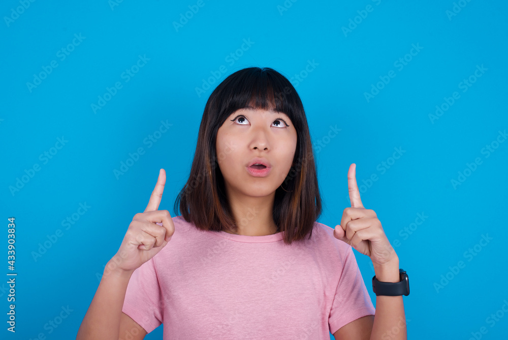 young beautiful asian woman wearing pink t-shirt against blue wall being amazed and surprised looking and pointing up with fingers showing something strange. young beautiful asian woman wearing pink t-shirt against blue wall being amazed and surprised looking and pointing up with fingers showing something strange.