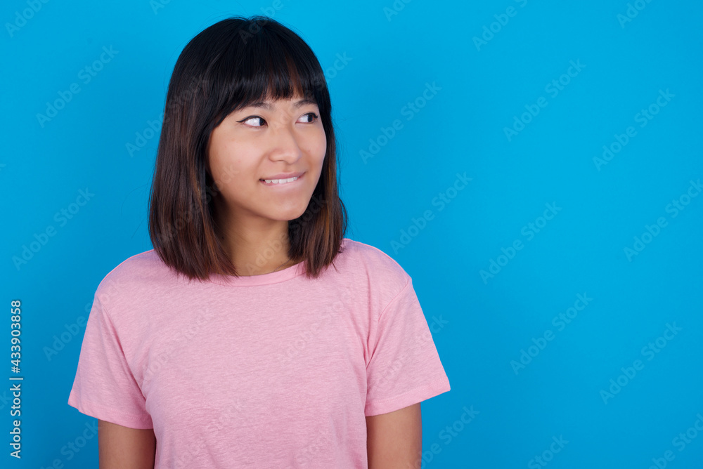 young beautiful asian woman wearing pink t-shirt against blue wall with thoughtful expression, looks away keeps hands down bitting his lip thinks about something pleasant. young beautiful asian woman wearing pink t-shirt against blue wall with thoughtful expression, looks away keeps hands down bitting his lip thinks about something pleasant.