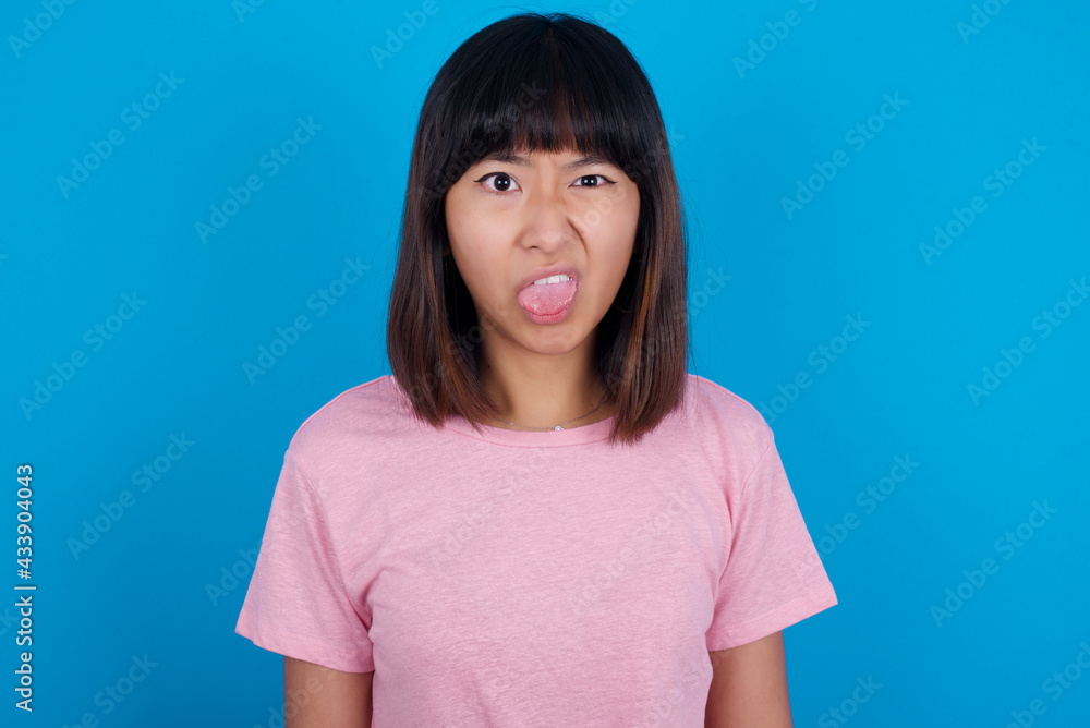 Body language. Disgusted stressed out young beautiful asian woman wearing pink t-shirt against blue wall, frowning face, demonstrating aversion to something. Body language. Disgusted stressed out young beautiful asian woman wearing pink t-shirt against blue wall, frowning face, demonstrating aversion to something.