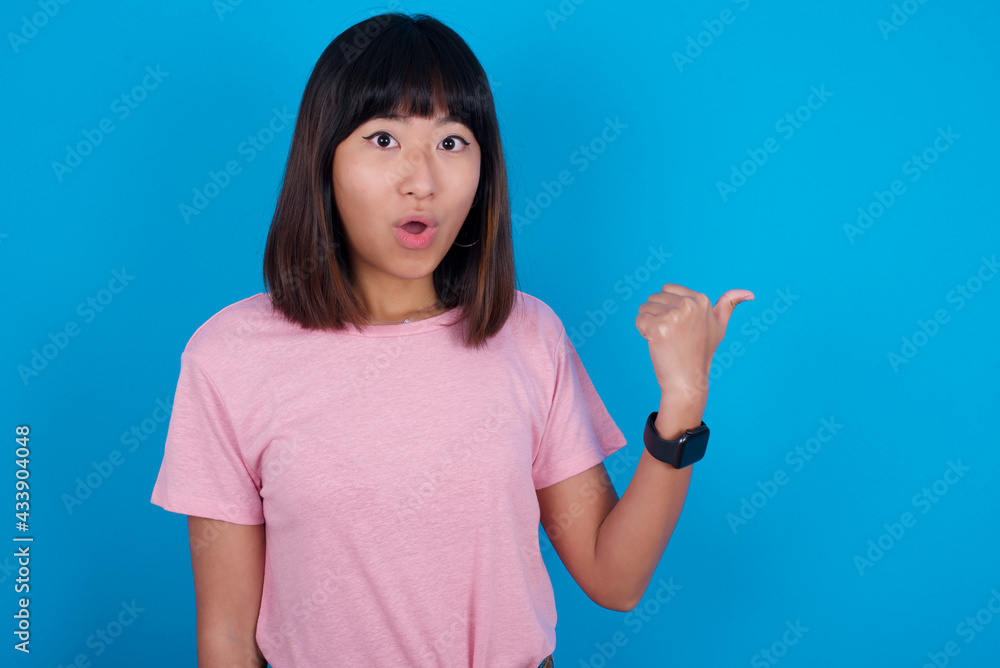 Shocked young beautiful asian woman wearing pink t-shirt against blue wall points with thumb away, indicates something. Check this out. Advertisement concept. Shocked young beautiful asian woman wearing pink t-shirt against blue wall points with thumb away, indicates something. Check this out. Advertisement concept.
