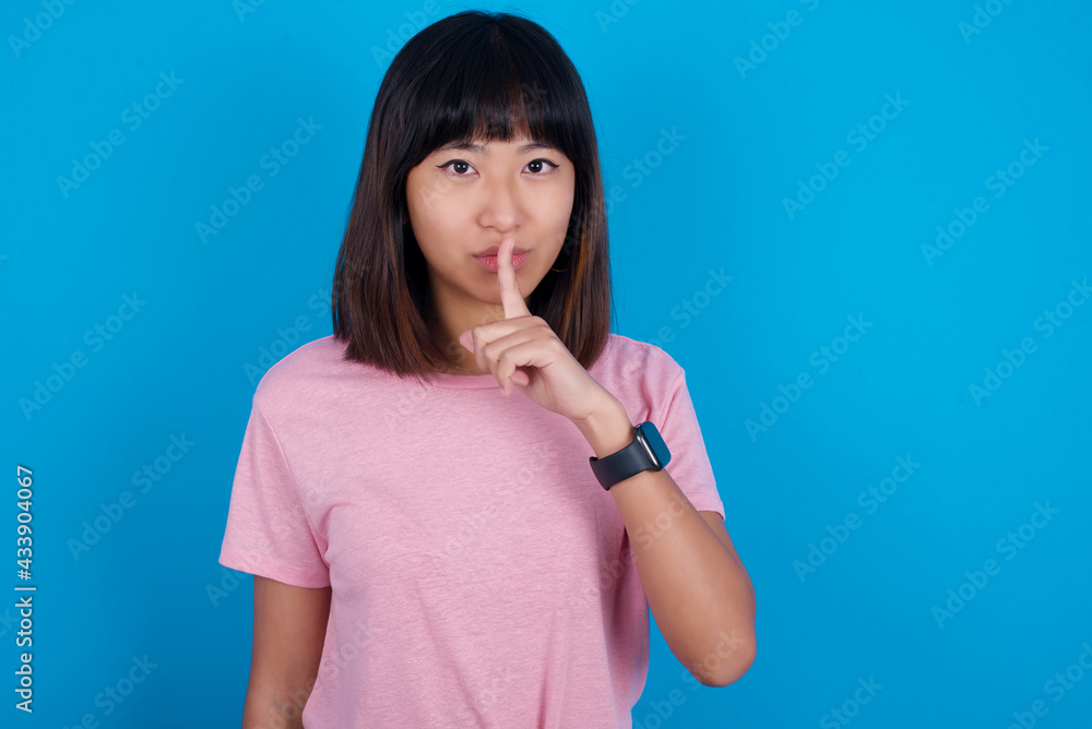 young beautiful asian woman wearing pink t-shirt against blue wall makes hush gesture, asks be quiet. Don't tell my secret or not speak too loud, please! young beautiful asian woman wearing pink t-shirt against blue wall makes hush gesture, asks be quiet. Don't tell my secret or not speak too loud, please!
