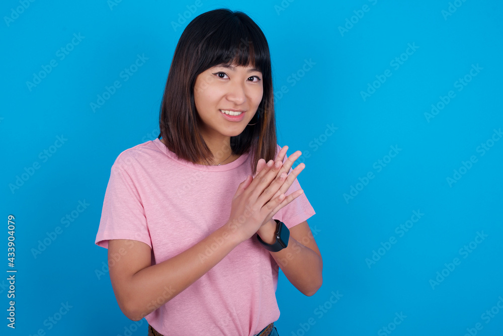 young beautiful asian woman wearing pink t-shirt against blue wall, feeling happy, smiling and clapping hands, saying congratulations with an applause. young beautiful asian woman wearing pink t-shirt against blue wall, feeling happy, smiling and clapping hands, saying congratulations with an applause.