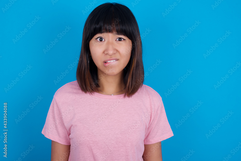 young beautiful asian woman wearing pink t-shirt against blue wall being nervous and scared biting lips looking camera with impatient expression, pensive. young beautiful asian woman wearing pink t-shirt against blue wall being nervous and scared biting lips looking camera with impatient expression, pensive.