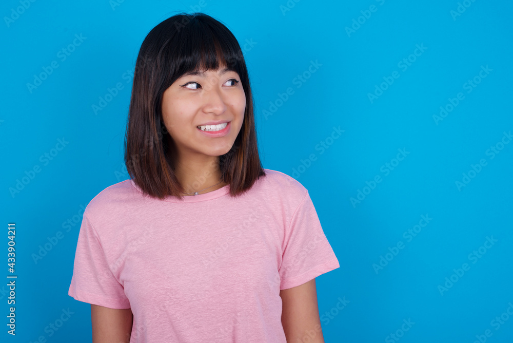 Oops! Portrait of young beautiful asian woman wearing pink t-shirt against blue wall clenches teeth and looks confusedly aside, realizes her bad mistake, Oops! Portrait of young beautiful asian woman wearing pink t-shirt against blue wall clenches teeth and looks confusedly aside, realizes her bad mistake,