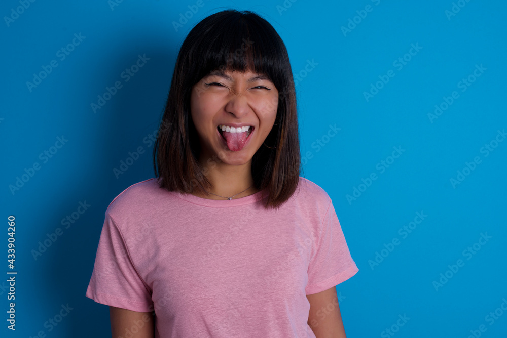 young beautiful asian woman wearing pink t-shirt against blue wall with happy and funny face smiling and showing tongue. young beautiful asian woman wearing pink t-shirt against blue wall with happy and funny face smiling and showing tongue.