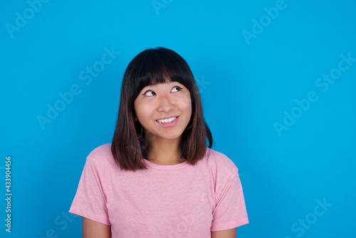 Portrait of mysterious young beautiful asian woman wearing pink t-shirt against blue wall looking up with enigmatic smile. Advertisement concept.