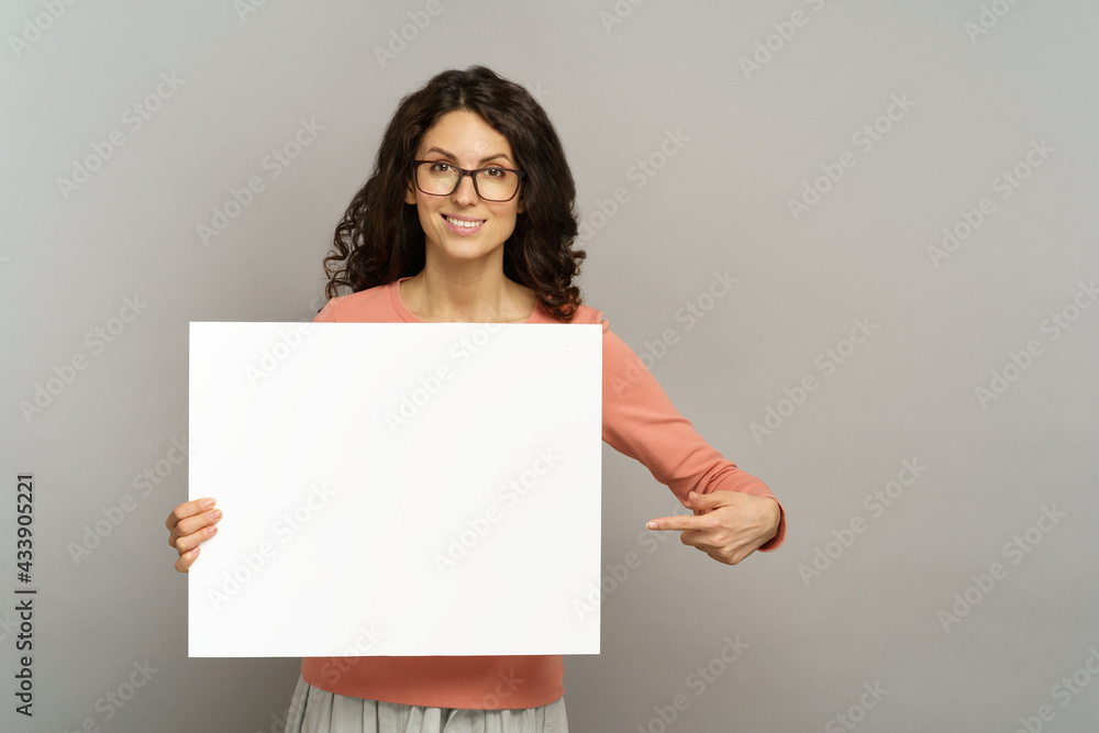 Smiling woman with empty poster pointing finger to white blank placard in hands, show advertisement on billboard. Attractive millennial businesswoman display profitable opportunity hold empty banner Smiling woman with empty poster pointing finger to white blank placard in hands, show advertisement on billboard. Attractive millennial businesswoman display profitable opportunity hold empty banner