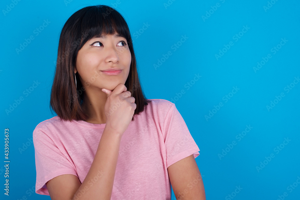 Young beautiful asian girl wearing pink t-shirt against blue background with hand under chin and looking sideways with doubtful and skeptical expression, suspect and doubt. Young beautiful asian girl wearing pink t-shirt against blue background with hand under chin and looking sideways with doubtful and skeptical expression, suspect and doubt.