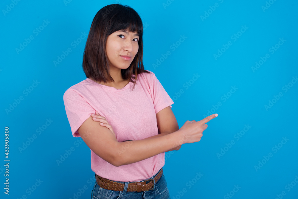 Young beautiful asian girl wearing pink t-shirt against blue background smiling broadly at camera, pointing fingers away, showing something interesting and exciting. Young beautiful asian girl wearing pink t-shirt against blue background smiling broadly at camera, pointing fingers away, showing something interesting and exciting.