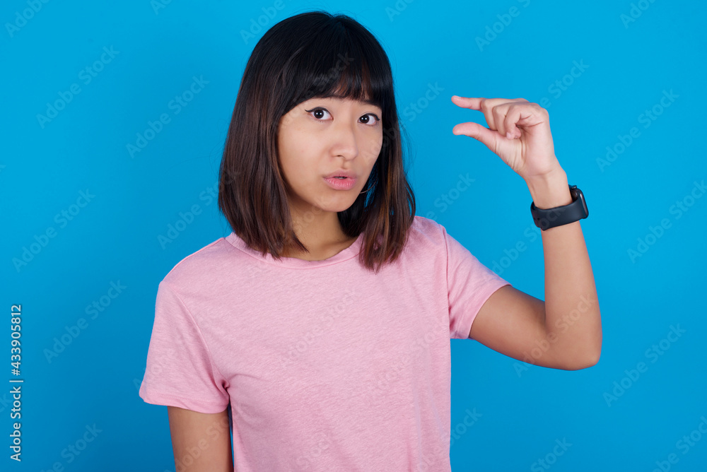 Shocked Young beautiful asian girl wearing pink t-shirt against blue background shows something little with hands, demonstrates size, opens mouth from surprise. Measurement concept. Shocked Young beautiful asian girl wearing pink t-shirt against blue background shows something little with hands, demonstrates size, opens mouth from surprise. Measurement concept.