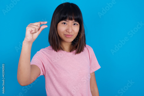 Upset Young beautiful asian girl wearing pink t-shirt against blue background shapes little gesture with hand demonstrates something very tiny small size. Not very much