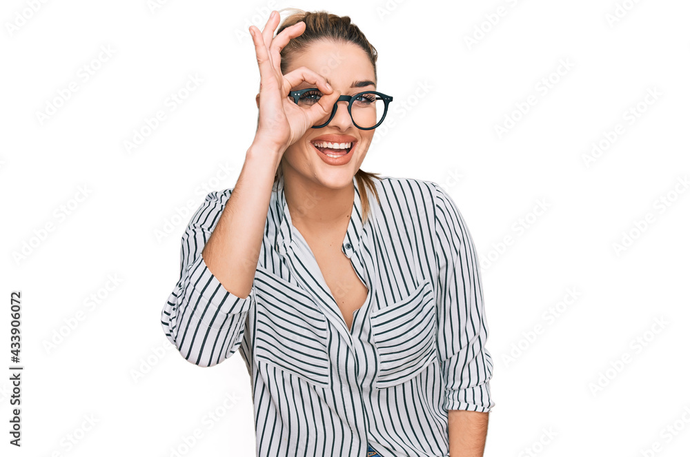 Young caucasian woman wearing business shirt and glasses doing ok gesture with hand smiling, eye looking through fingers with happy face. Young caucasian woman wearing business shirt and glasses doing ok gesture with hand smiling, eye looking through fingers with happy face.