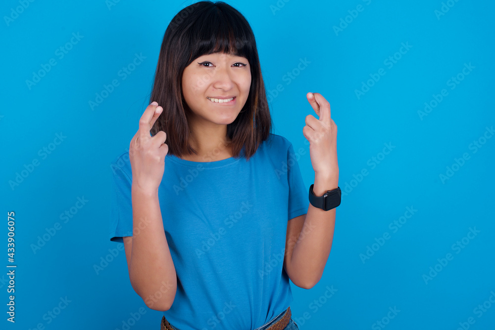 Young beautiful asian girl wearing blue t-shirt against blue background holding fingers crossed with worried expression hoping boss didn't noticed mistakes at work. Young beautiful asian girl wearing blue t-shirt against blue background holding fingers crossed with worried expression hoping boss didn't noticed mistakes at work.
