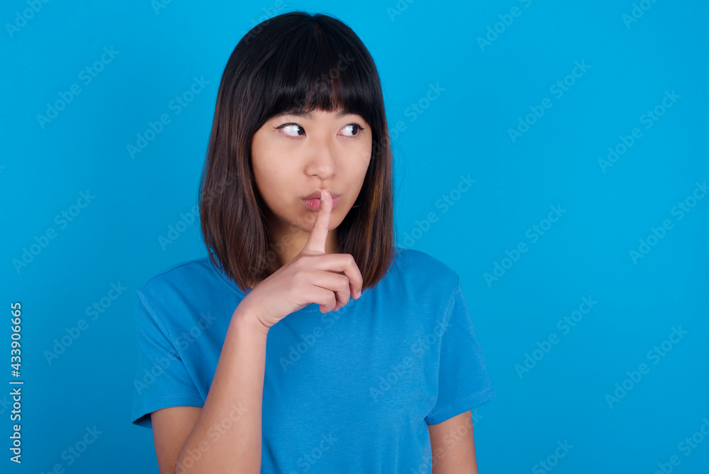 young beautiful asian girl wearing blue t-shirt against blue background makes silence gesture, keeps index finger to lips makes hush sign. Asks not to share secret. young beautiful asian girl wearing blue t-shirt against blue background makes silence gesture, keeps index finger to lips makes hush sign. Asks not to share secret.