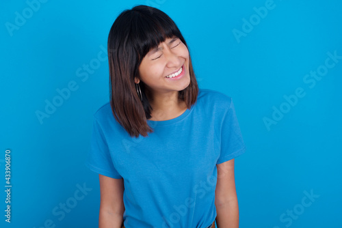 Positive young beautiful asian girl wearing blue t-shirt against blue background with overjoyed expression closes eyes and laughs shows white perfect teeth © Jihan
