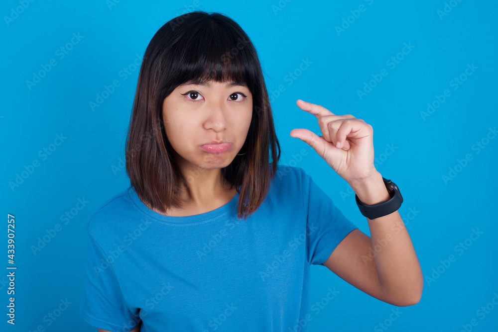 young beautiful asian girl wearing blue t-shirt against blue background purses lip and gestures with hand, shows something very little. young beautiful asian girl wearing blue t-shirt against blue background purses lip and gestures with hand, shows something very little.
