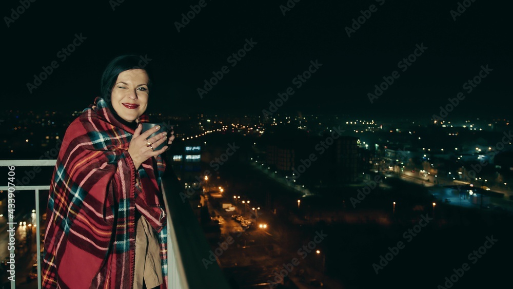 Portrait of a woman on the terrace of a high-rise building on a background of night city and blurred lights. Woman drinking tea or coffee wrapped in a warm blanket.