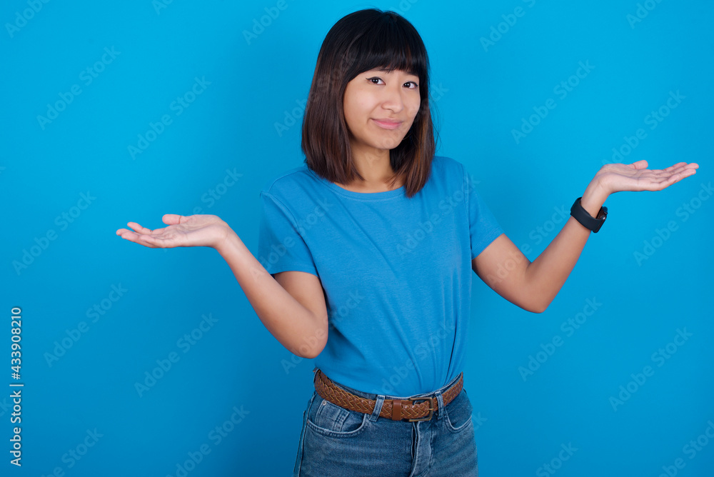 Careless attractive young beautiful asian girl wearing blue t-shirt against blue background shrugging shoulders, oops. Careless attractive young beautiful asian girl wearing blue t-shirt against blue background shrugging shoulders, oops.