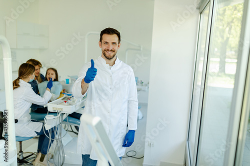 Male dentist standing in dental office, wearing gloves and white coat, showing thumbs up.