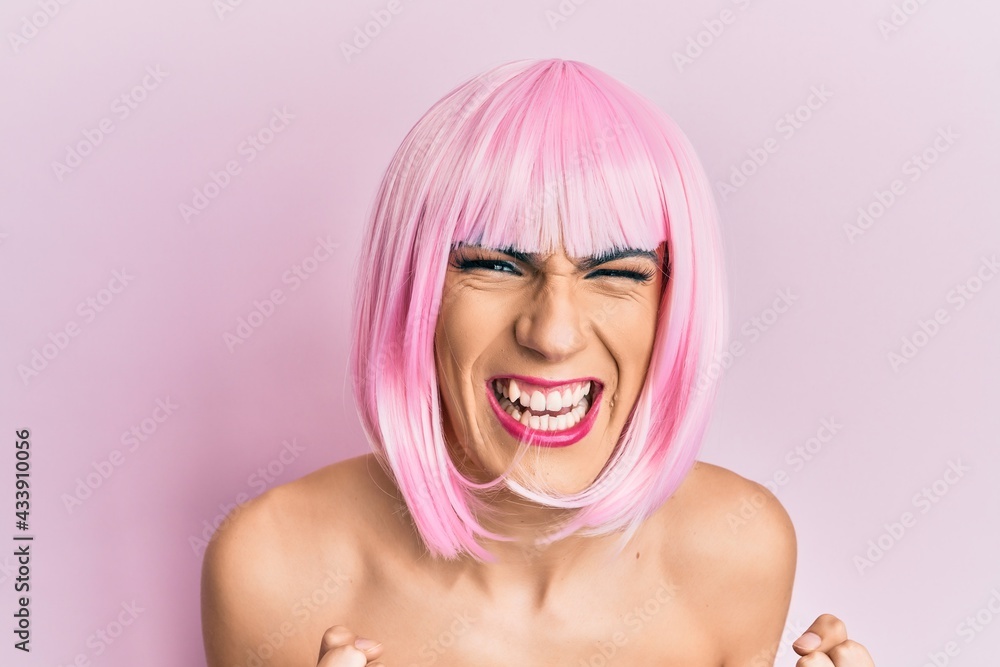 Young man wearing woman make up wearing pink wig very happy and excited doing winner gesture with arms raised, smiling and screaming for success. celebration concept. Young man wearing woman make up wearing pink wig very happy and excited doing winner gesture with arms raised, smiling and screaming for success. celebration concept.