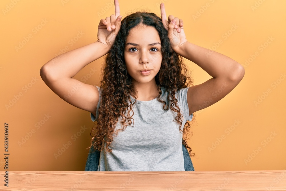 Teenager hispanic girl wearing casual clothes sitting on the table doing funny gesture with finger over head as bull horns Teenager hispanic girl wearing casual clothes sitting on the table doing funny gesture with finger over head as bull horns