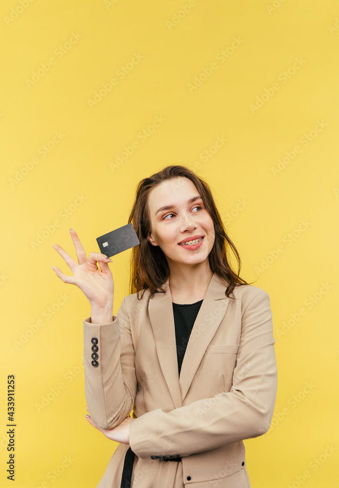Cute positive lady in a beige jacket poses with a bank card on a yellow background and looks away with a smile on her face. Cute positive lady in a beige jacket poses with a bank card on a yellow background and looks away with a smile on her face.
