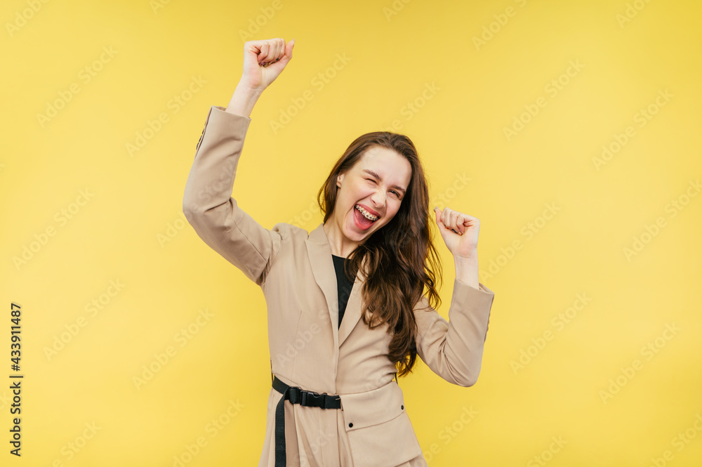 Joyful attractive woman in a beige suit emotionally celebrates on a yellow background, dances with happiness with raised hands and looks at the camera. Joyful attractive woman in a beige suit emotionally celebrates on a yellow background, dances with happiness with raised hands and looks at the camera.