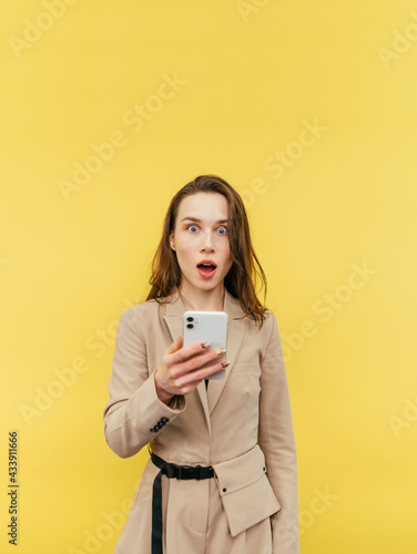 Portrait of a shocked lady in a suit with a smartphone in his hands on a yellow background, looking at the camera with his mouth open in surprise. Vertical.