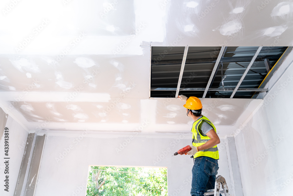 Asian construction worker assemble a suspended ceiling with drywall and fixing the drywall to the ceiling metal frame with screwdriver. Renovation, construction and ceiling installation concept. - obrazy, fototapety, plakaty 