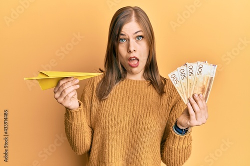 Beautiful caucasian woman holding paper airplane and 500 norwegian krone in shock face, looking skeptical and sarcastic, surprised with open mouth