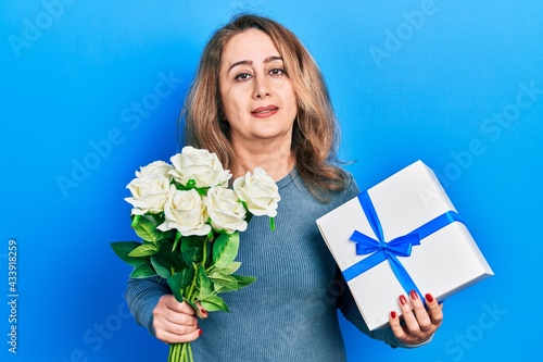 Middle age caucasian woman holding anniversary present and bouquet of flowers relaxed with serious expression on face. simple and natural looking at the camera.