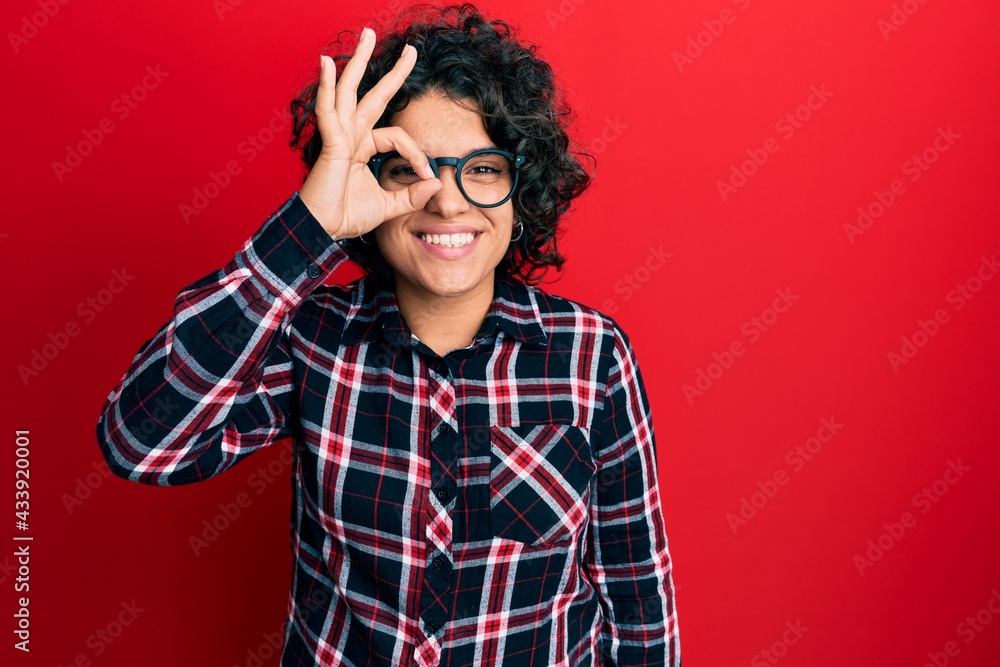 Young hispanic woman with curly hair wearing casual clothes and glasses doing ok gesture with hand smiling, eye looking through fingers with happy face. Young hispanic woman with curly hair wearing casual clothes and glasses doing ok gesture with hand smiling, eye looking through fingers with happy face.