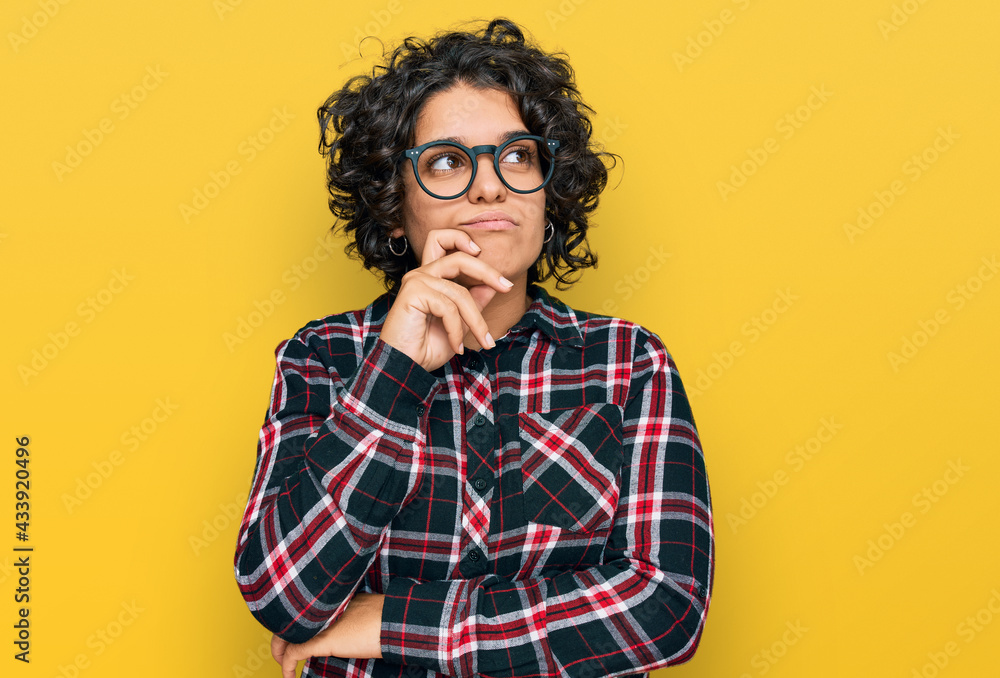 Young hispanic woman with curly hair wearing casual clothes and glasses serious face thinking about question with hand on chin, thoughtful about confusing idea Young hispanic woman with curly hair wearing casual clothes and glasses serious face thinking about question with hand on chin, thoughtful about confusing idea