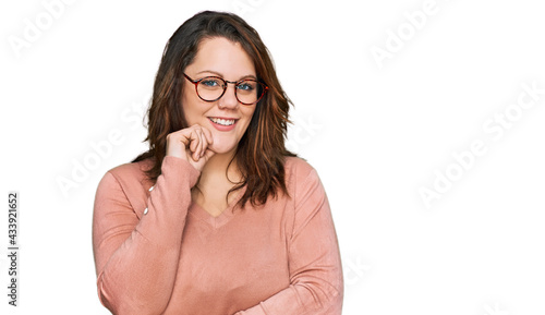 Young plus size woman wearing casual clothes and glasses looking confident at the camera smiling with crossed arms and hand raised on chin. thinking positive.