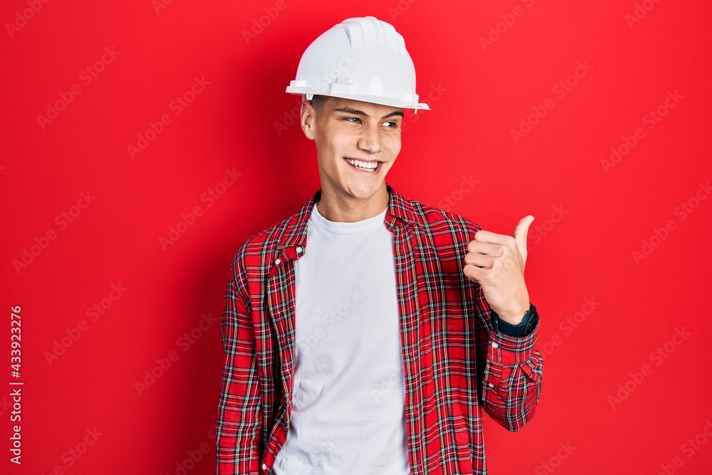 Young hispanic man wearing architect hardhat smiling with happy face looking and pointing to the side with thumb up.