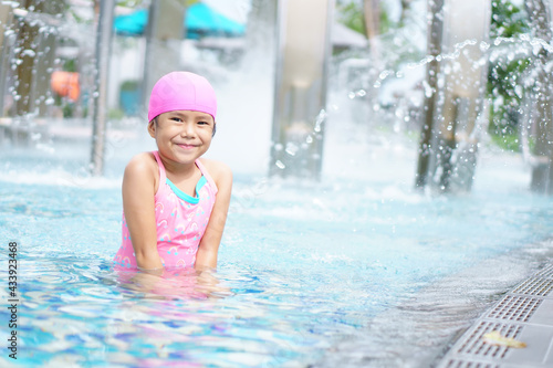 Asian child cute or kid girl wearing swimsuit with cap on swimming pool and enjoy smiling with happy fun to cold in water park fountain for play and refreshing or relax with exercise on summer holiday © kornnphoto