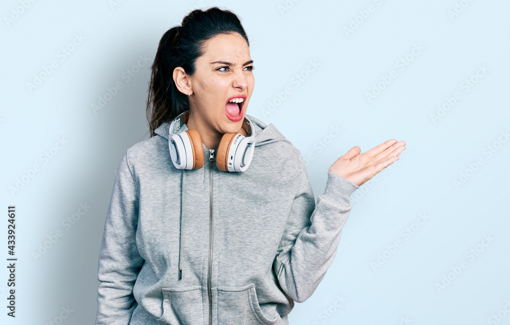 Young hispanic woman wearing gym clothes, using headphones and smart watch angry and mad screaming frustrated and furious, shouting with anger. rage and aggressive concept. Young hispanic woman wearing gym clothes, using headphones and smart watch angry and mad screaming frustrated and furious, shouting with anger. rage and aggressive concept.
