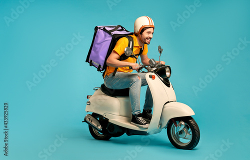 Young courier, delivery man in uniform with thermo backpack on a moped isolated on blue background. Fast transport express home delivery. Online order.