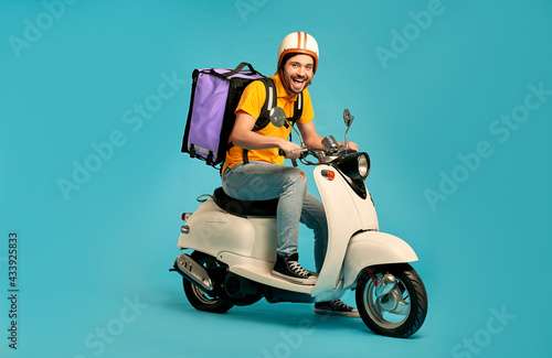 Fotografie Young courier, delivery man in uniform with thermo backpack on a moped isolated on blue background