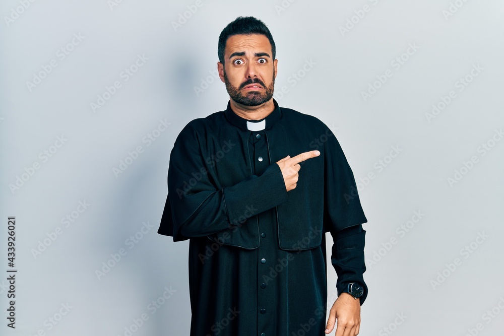 Obraz premium Handsome hispanic man with beard wearing catholic priest robe pointing aside worried and nervous with forefinger, concerned and surprised expression