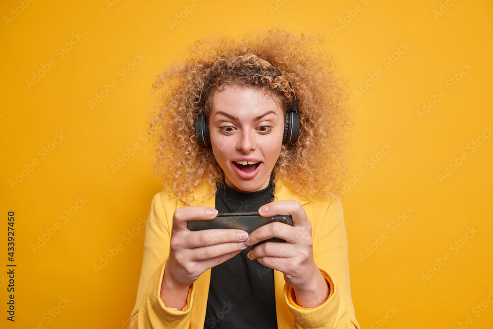 Indoor shot of surprised cheerful curly haired woman concentrated at smartphone display while playing video games uses wireless stereo headphones enjoys spare time isolated over yellow background Indoor shot of surprised cheerful curly haired woman concentrated at smartphone display while playing video games uses wireless stereo headphones enjoys spare time isolated over yellow background