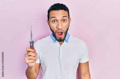 Hispanic man with beard holding pocket knife scared and amazed with open mouth for surprise, disbelief face